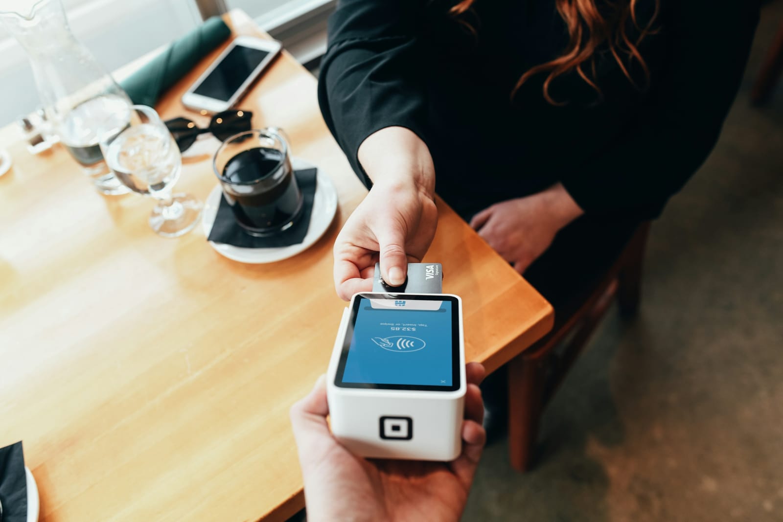 A customer paying by card at a cafe table, inserting a Visa card into a handheld payment terminal showing a $32.85 total.