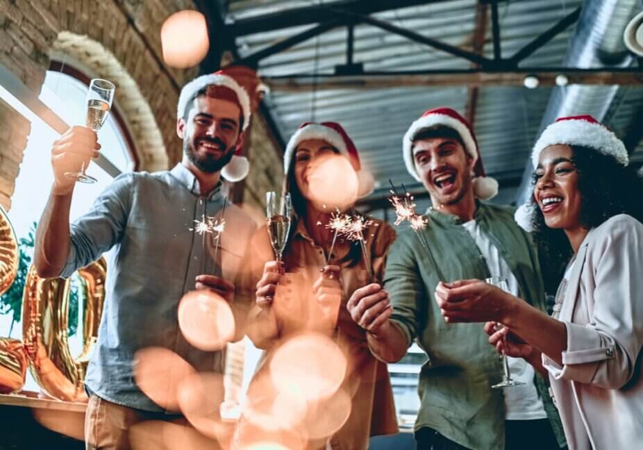Merry Christmas and Happy New Year 2020!Multiracial young creative people are celebrating holiday in modern office. Group of young business people are drinking champagne with sparkling bengal lights in coworking. Successful hipster team of freelancers.