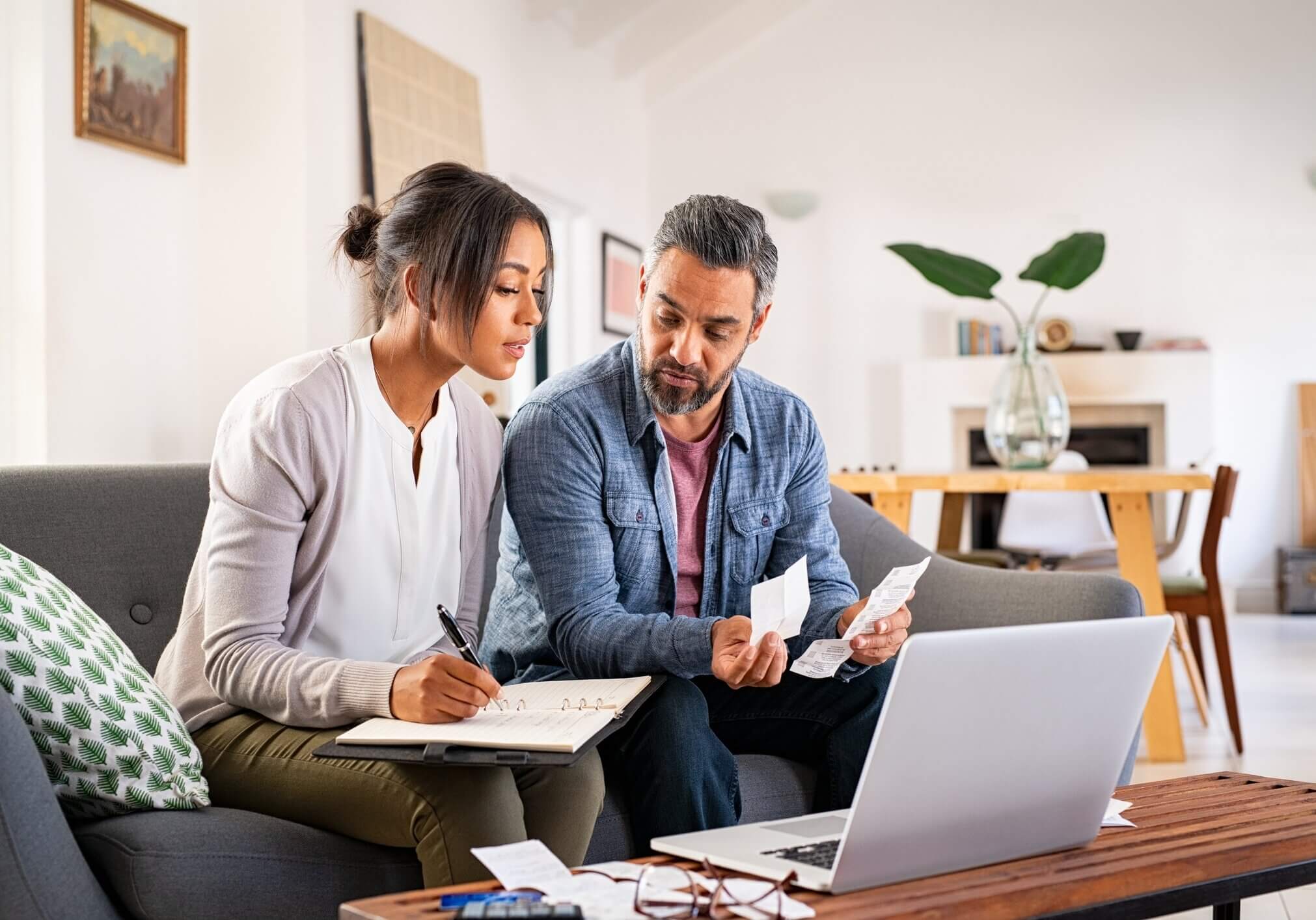 Mature couple calculating bills at home using laptop and calculator. Multiethnic couple working on computer while calculating finances sitting on couch. Mature indian man with african american woman at home analyzing their finance with documents.