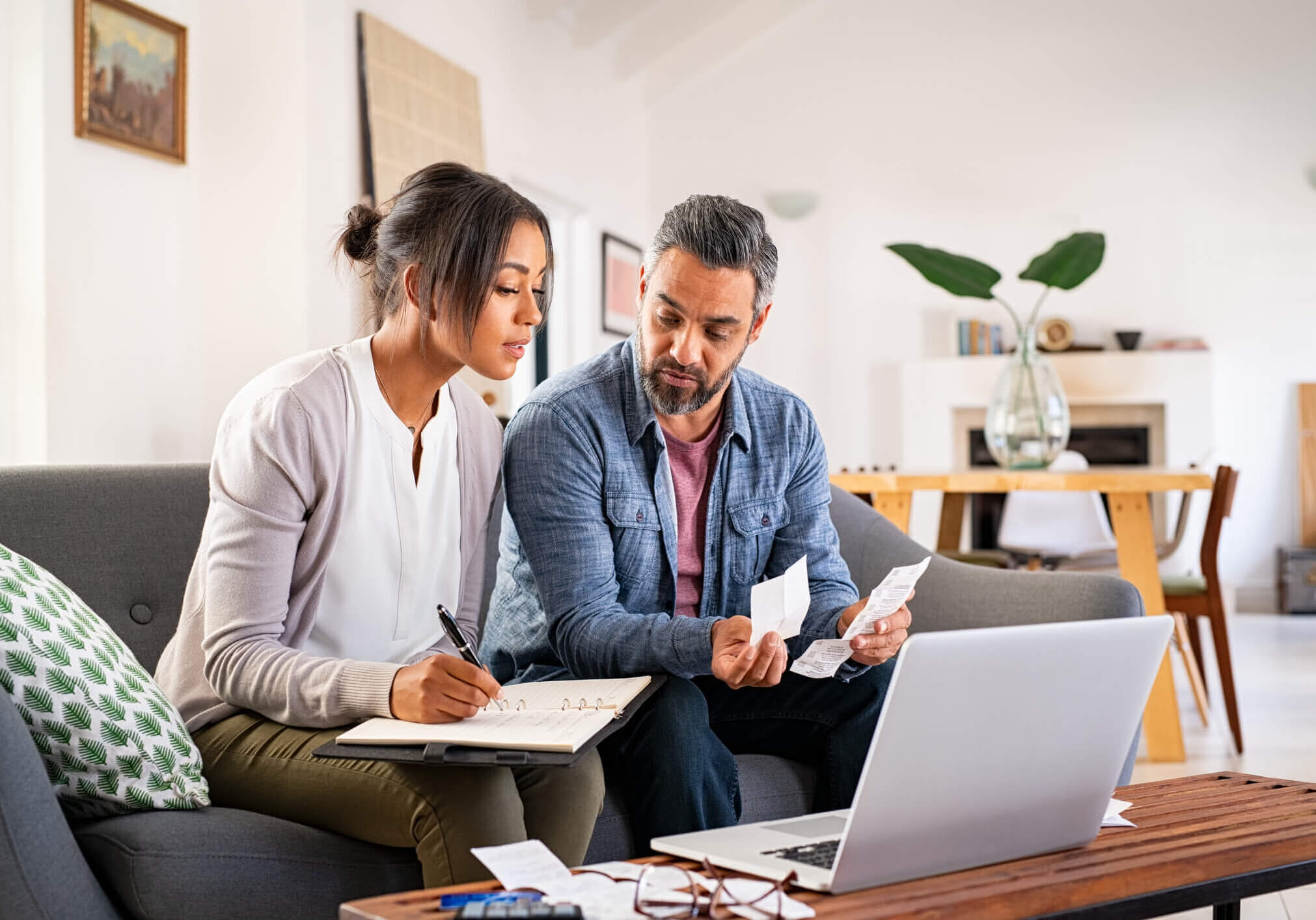 Mature couple calculating bills at home using laptop and calculator. Multiethnic couple working on computer while calculating finances sitting on couch. Mature indian man with african american woman at home analyzing their finance with documents.