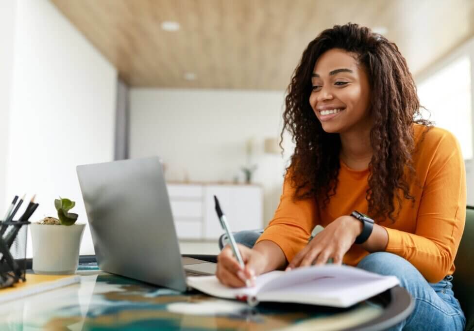 Note Taking Concept. Smiling young African American woman sitting at desk working on laptop and writing letter in paper notebook, holding pen and looking at screen. Happy millennial female using pc